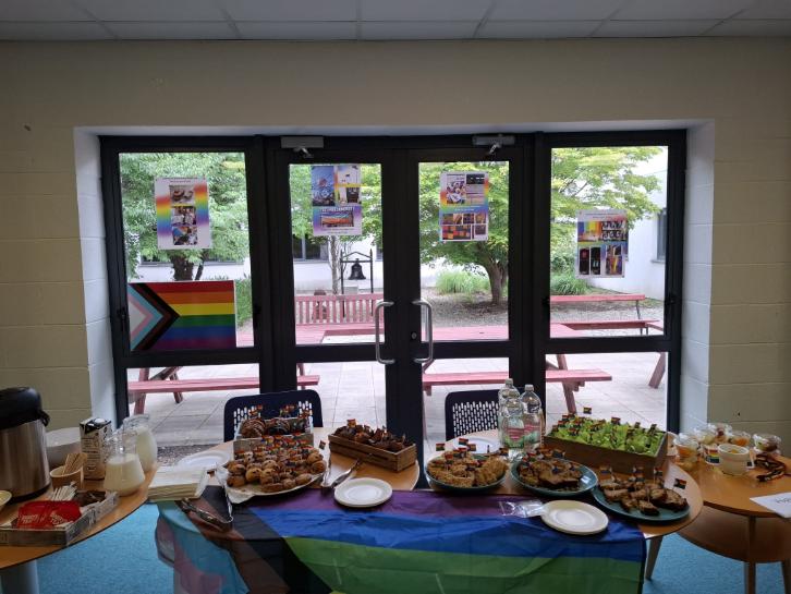 A table set up for a Pride-themed coffee morning at the UCD School of Veterinary Medicine, decorated with a Progress Pride flag and rainbow posters on the windows. Plates of pastries, cakes, and sandwiches are arranged on tables with coffee, milk, and water available.